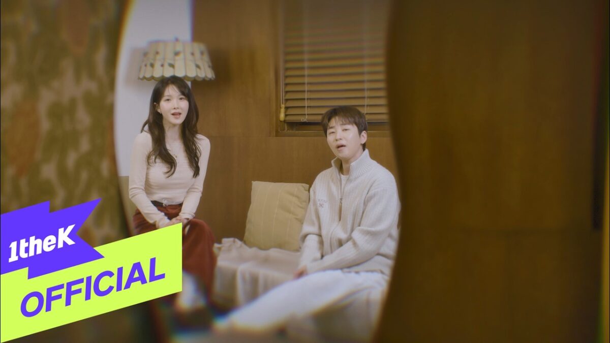 Dindin e Cho Hyun Young sentados em um sofá, cantando no clipe "Always You", com iluminação suave e ambiente aconchegante.