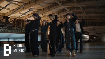 Grupo BTS performando a coreografia da música "SWIM" em um ambiente amplo e iluminado, com aviões ao fundo.