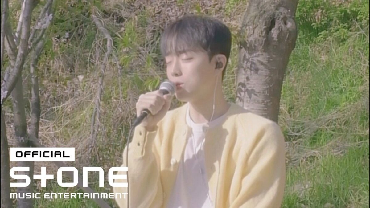 Cantor em um suéter amarelo canta em um ambiente natural, com árvores ao fundo e microfone na mão.