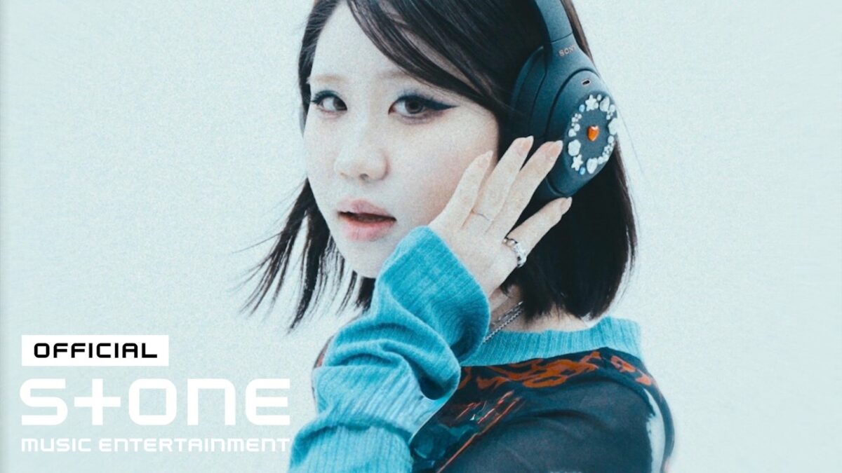 Cantora com fones de ouvido, usando um suéter azul, olhando diretamente para a câmera em fundo claro.