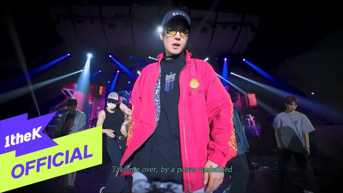 Artista em destaque com jaqueta rosa performando em palco iluminado, rodeado por dançarinos.