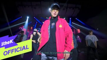 Artista em destaque com jaqueta rosa performando em palco iluminado, rodeado por dançarinos.