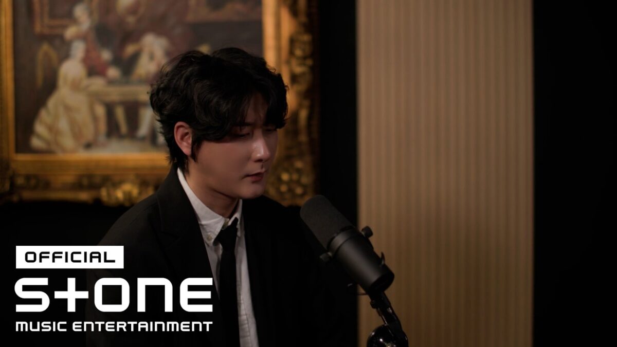 Jungsuk canta em um estúdio elegante, com um microfone à sua frente e uma pintura clássica ao fundo.