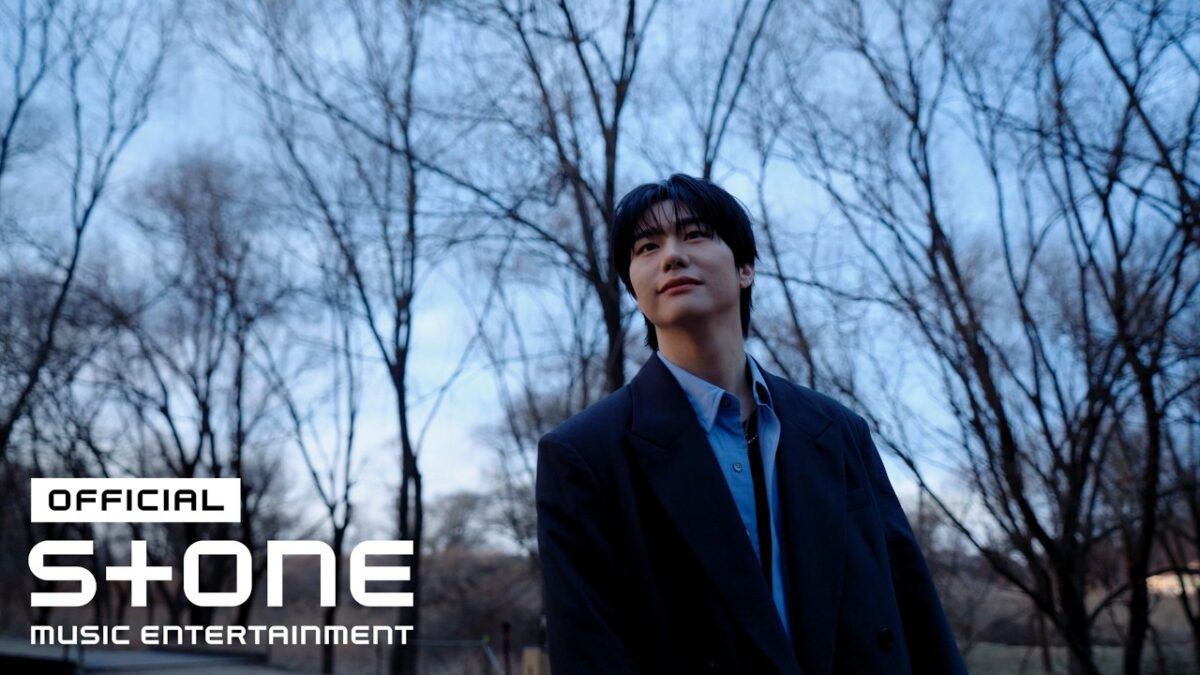 Kang Seungsik sorri enquanto observa o céu em um ambiente natural com árvores secas ao fundo.