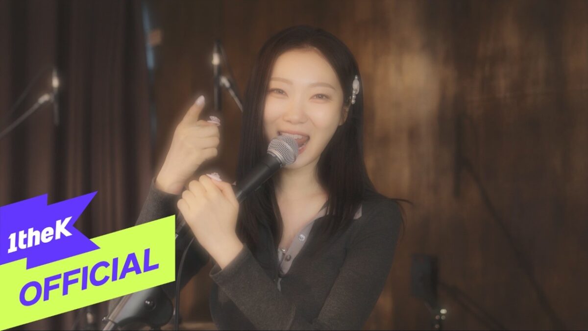 Sohlhee canta animada com microfone em mãos, sorrindo para a câmera em um cenário musical.