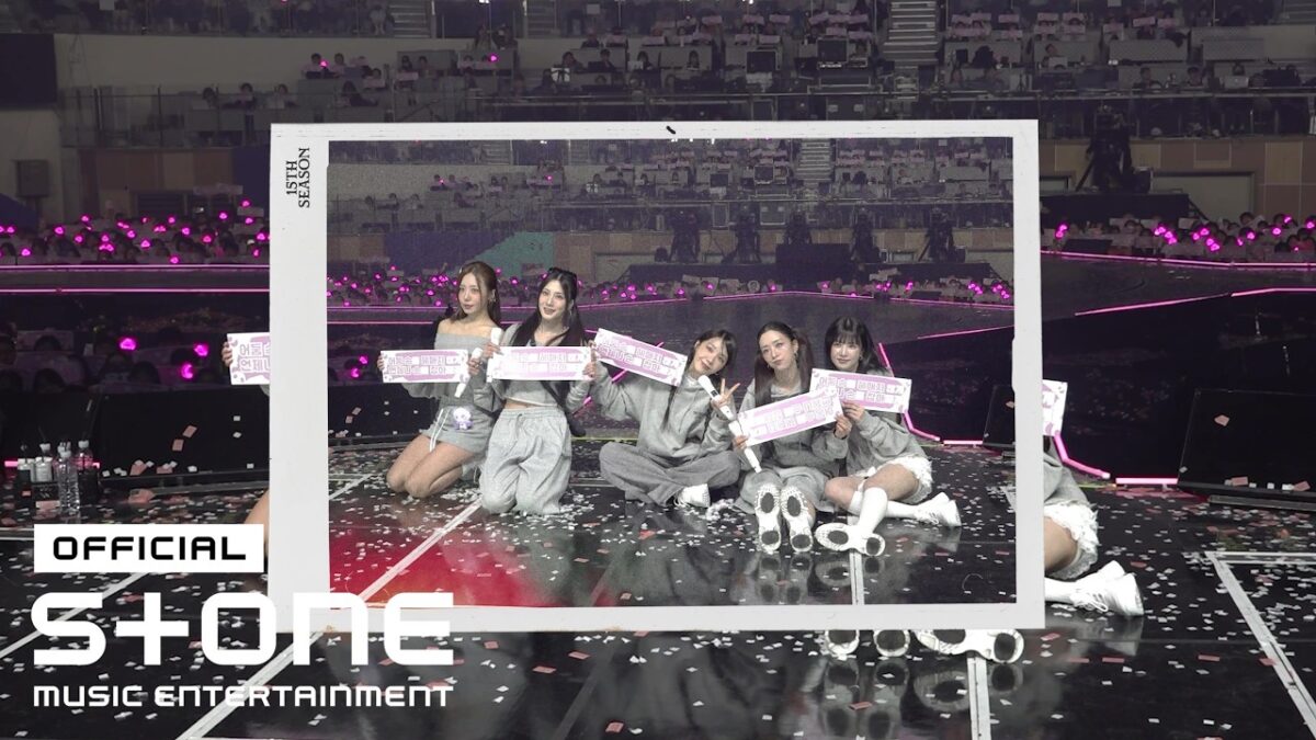 Cinco integrantes do Apink posam em um palco com confetes, segurando cartazes e cercadas por fãs com luzes rosas.
