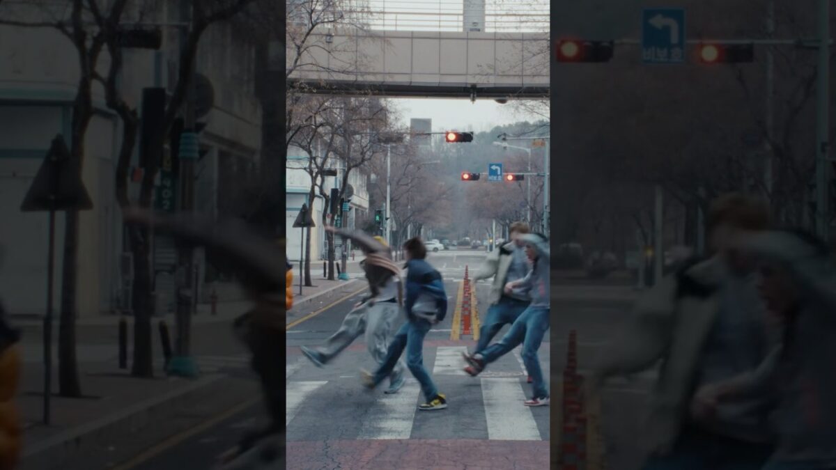 Grupo de pessoas atravessando uma rua em movimento, com semáforo vermelho ao fundo e árvores sem folhas nas laterais.