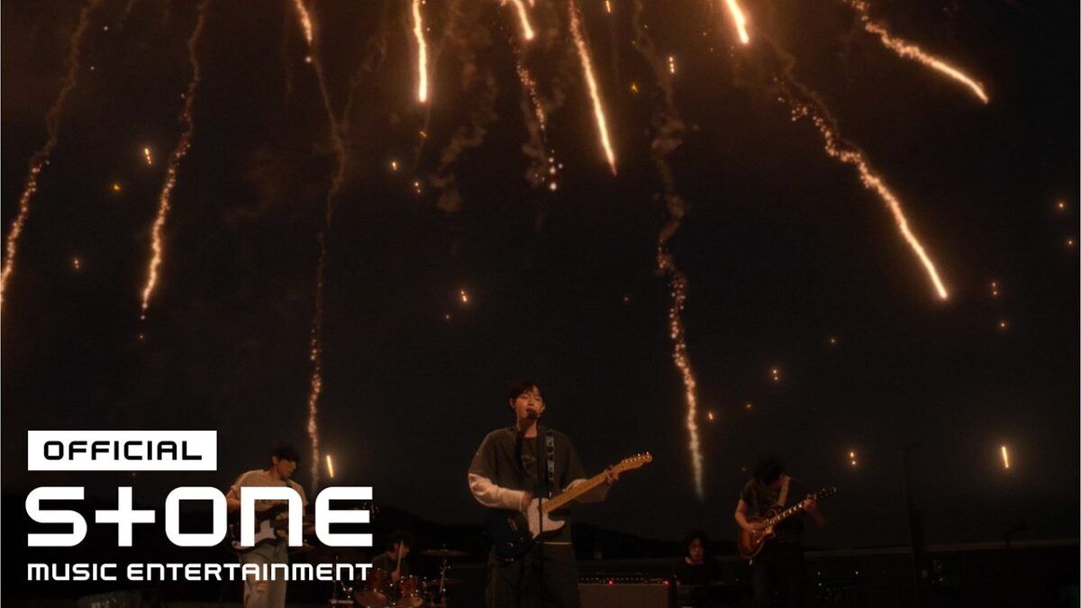 Kim Jae Hwan se apresenta com fogos de artifício iluminando o céu ao fundo.