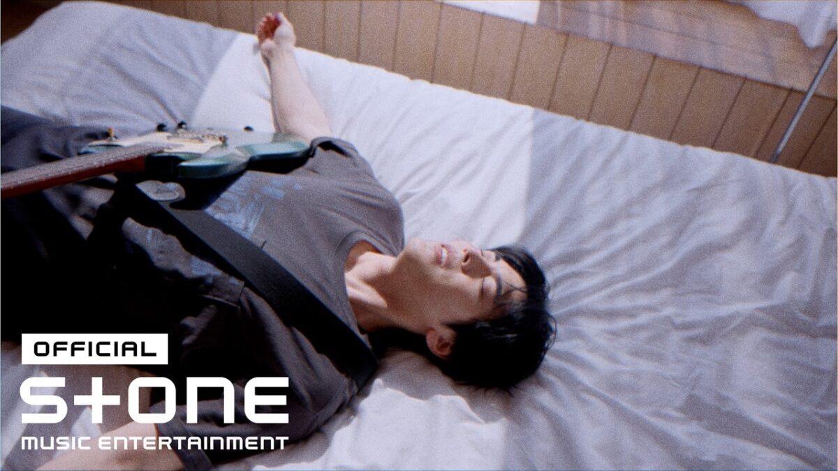Homem deitado em uma cama, sorrindo, com uma guitarra ao seu lado e luz natural entrando pela janela.