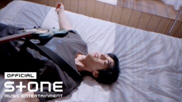 Homem deitado em uma cama, sorrindo, com uma guitarra ao seu lado e luz natural entrando pela janela.