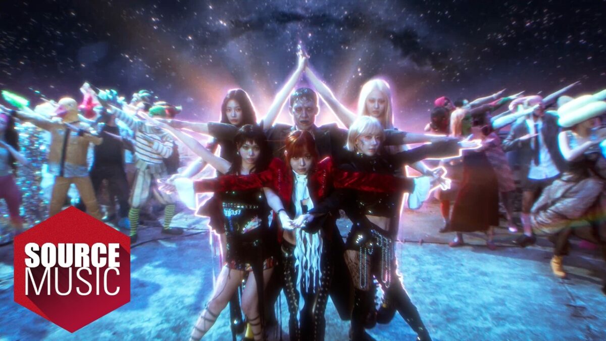 Grupo de artistas em trajes vibrantes, posando em um cenário cósmico iluminado, com expressões de celebração.