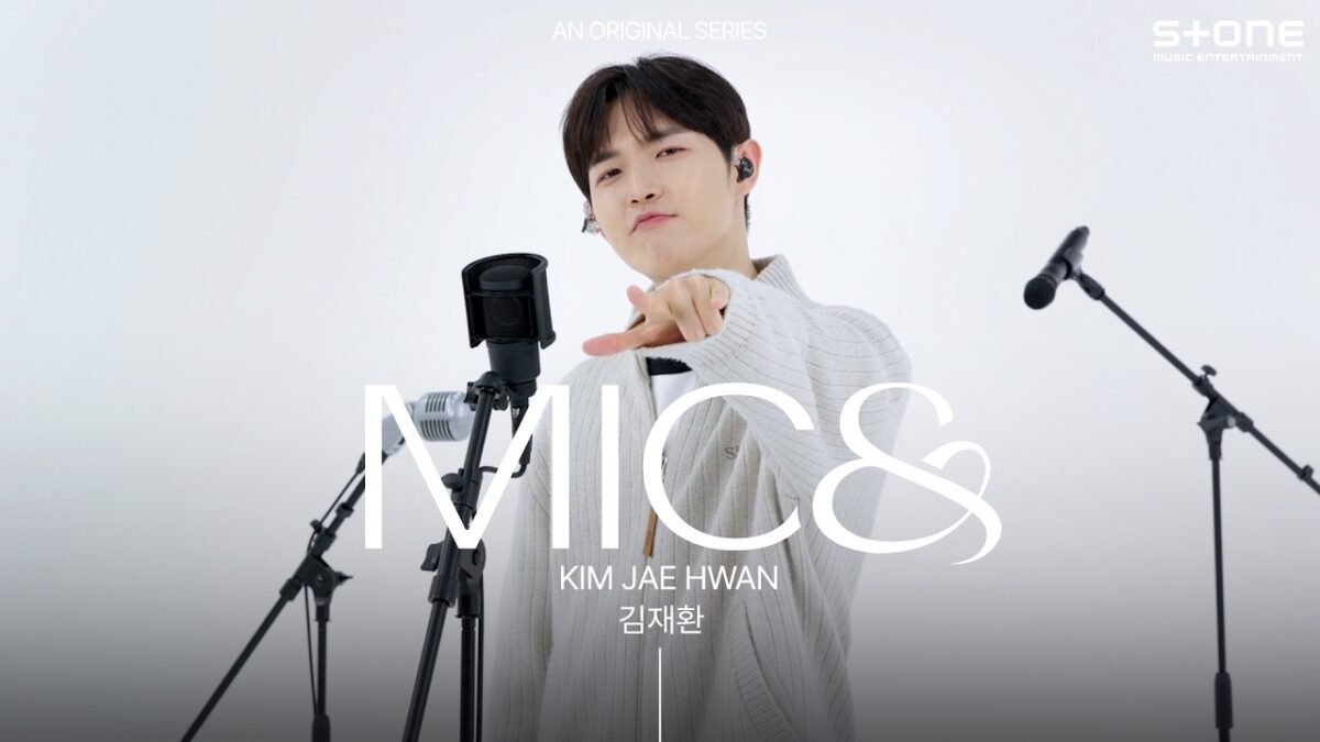 Kim Jae-hwan aponta para a câmera durante sua apresentação ao vivo, cercado por microfones em um cenário minimalista.