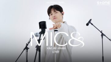 Kim Jae-hwan aponta para a câmera durante sua apresentação ao vivo, cercado por microfones em um cenário minimalista.