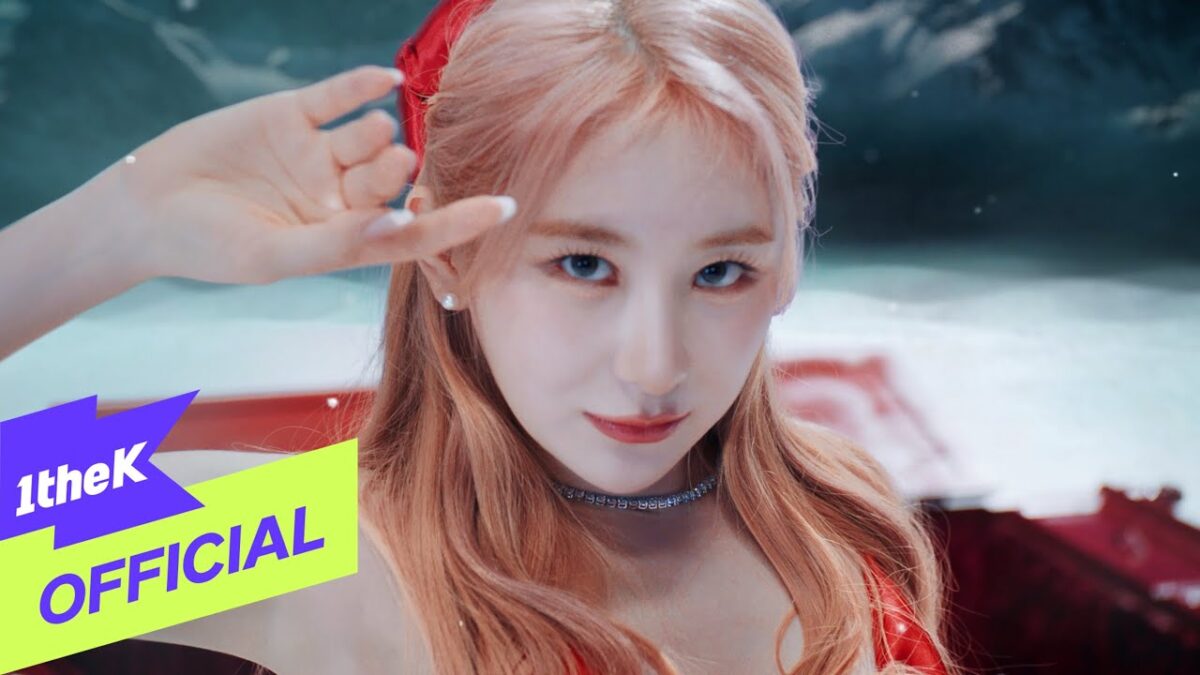 Lee Chae Yeon com cabelo longo e rosa, usando um vestido vermelho, faz um gesto de saudação em cenário vibrante.