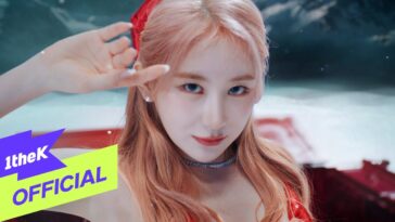 Lee Chae Yeon com cabelo longo e rosa, usando um vestido vermelho, faz um gesto de saudação em cenário vibrante.