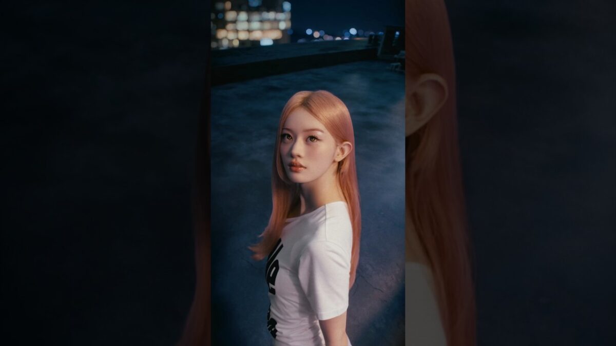 Jovem com cabelo longo e rosa, vestindo camiseta branca, posa em um telhado à noite com luzes da cidade ao fundo.