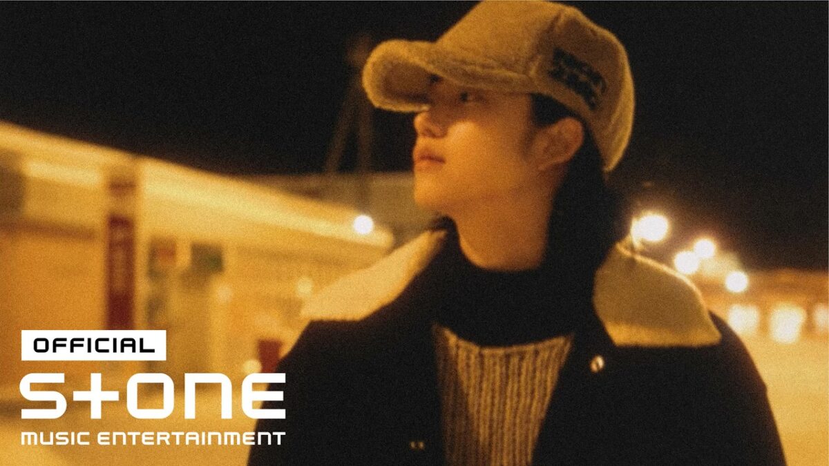 Jovem com boné e casaco escuro observa o ambiente iluminado à noite, transmitindo uma atmosfera contemplativa.