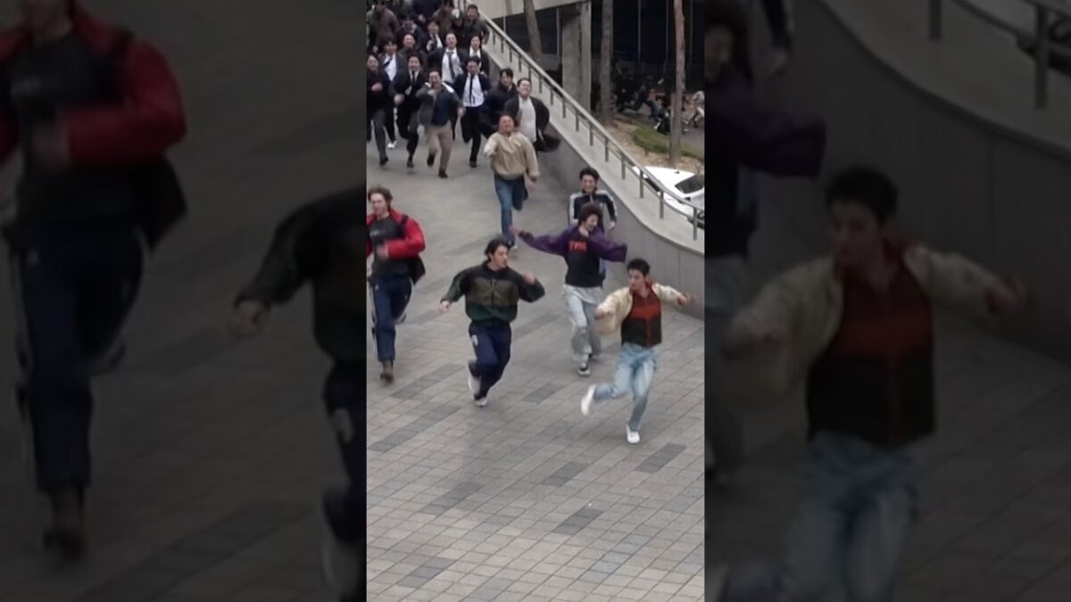 Grupo de jovens correndo em um espaço urbano, cercados por pessoas em trajes formais.