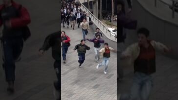 Grupo de jovens correndo em um espaço urbano, cercados por pessoas em trajes formais.