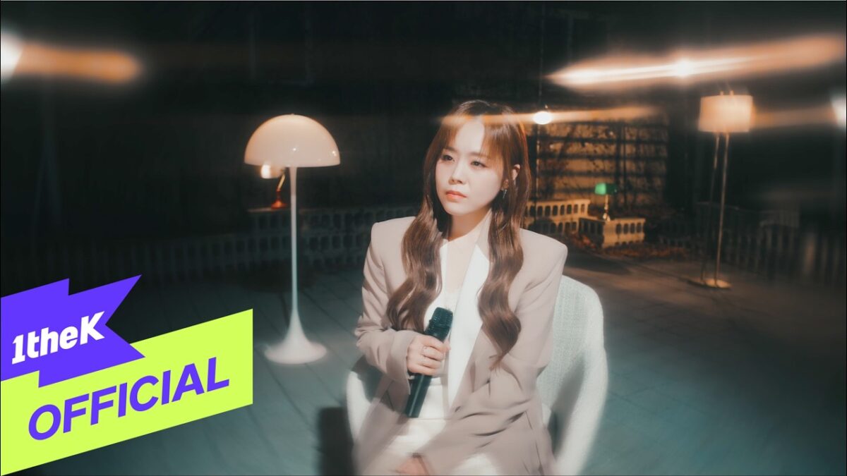 Lee Ye Joon sentada em uma cadeira, segurando um microfone, em um ambiente iluminado com uma lâmpada ao fundo.