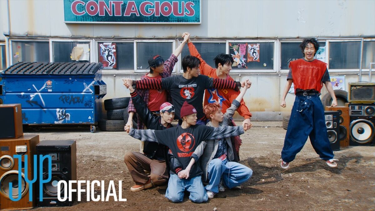 Sete jovens dançam em uma coreografia animada, cercados por equipamentos de som e um ambiente urbano vibrante.