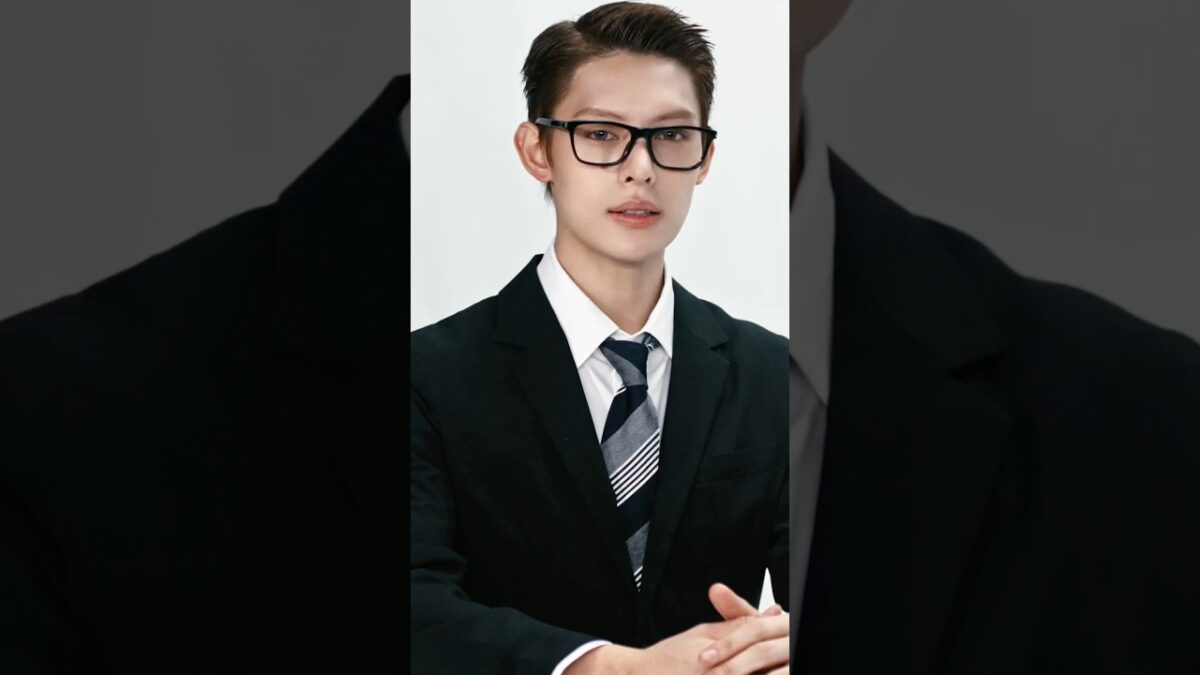 Jovem de terno e gravata, usando óculos, com expressão confiante, sentado em uma mesa.