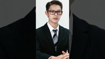 Jovem de terno e gravata, usando óculos, com expressão confiante, sentado em uma mesa.