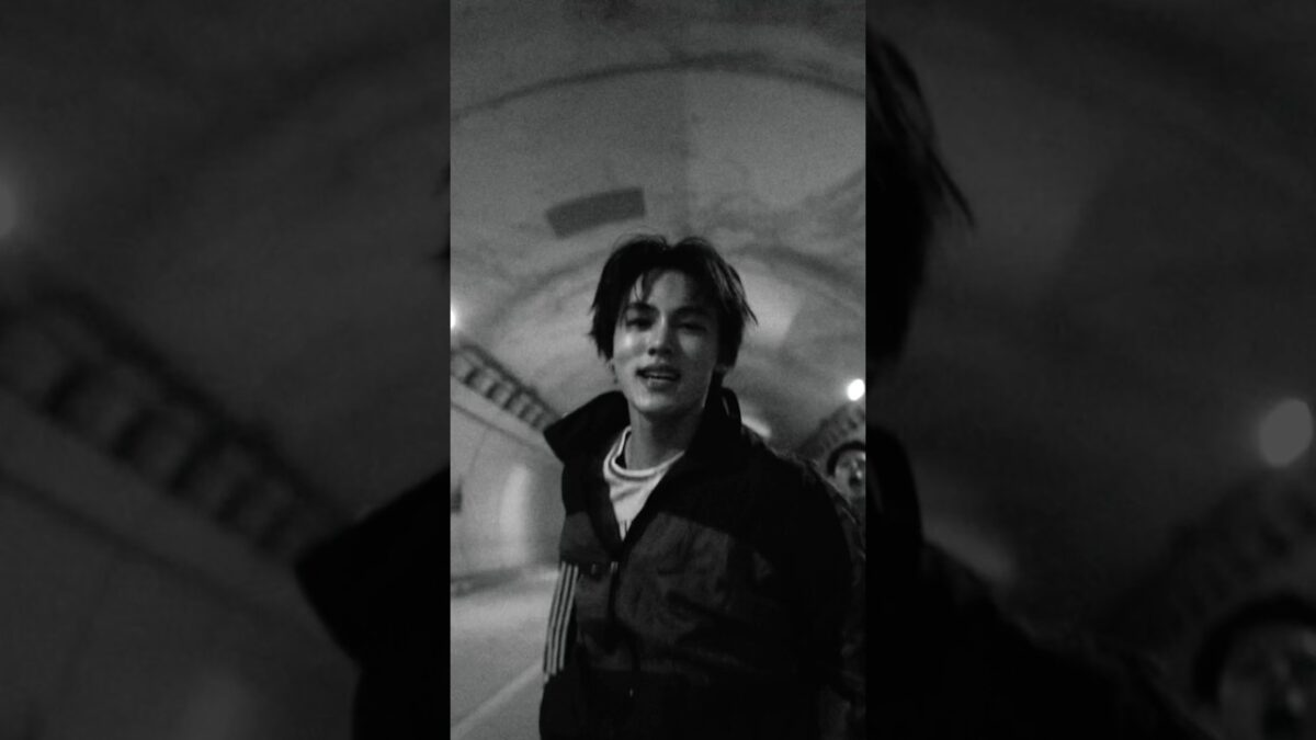 Jovem sorridente em túnel, vestindo jaqueta escura, com luzes ao fundo em preto e branco.