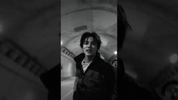 Jovem sorridente em túnel, vestindo jaqueta escura, com luzes ao fundo em preto e branco.