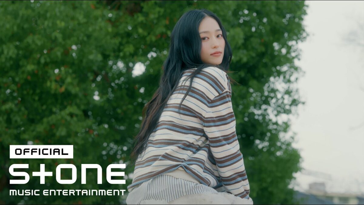 Yeeun, com cabelo longo e liso, usa uma blusa listrada enquanto olha por cima do ombro em um cenário verde.