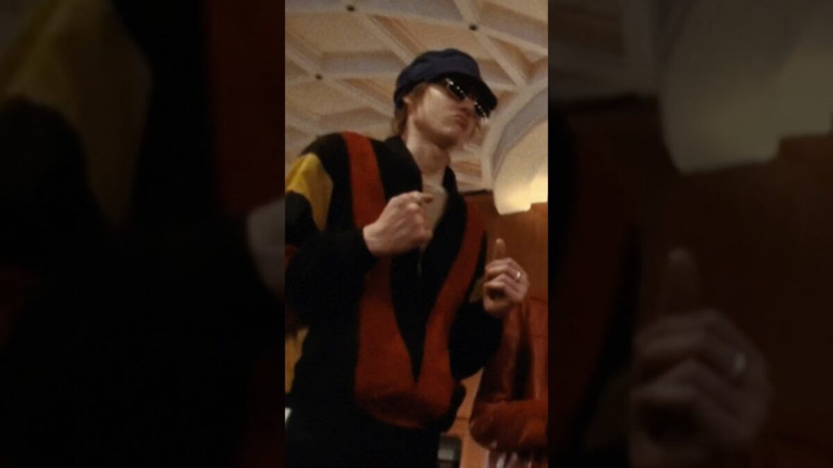 Artista performa com roupas coloridas e óculos escuros em ambiente interno, transmitindo energia vibrante.