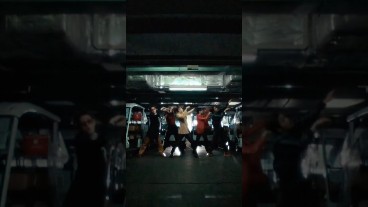 Grupo de dançarinos se apresenta em um ambiente escuro, com iluminação suave e veículos ao fundo.
