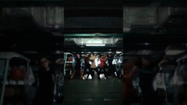 Grupo de dançarinos se apresenta em um ambiente escuro, com iluminação suave e veículos ao fundo.