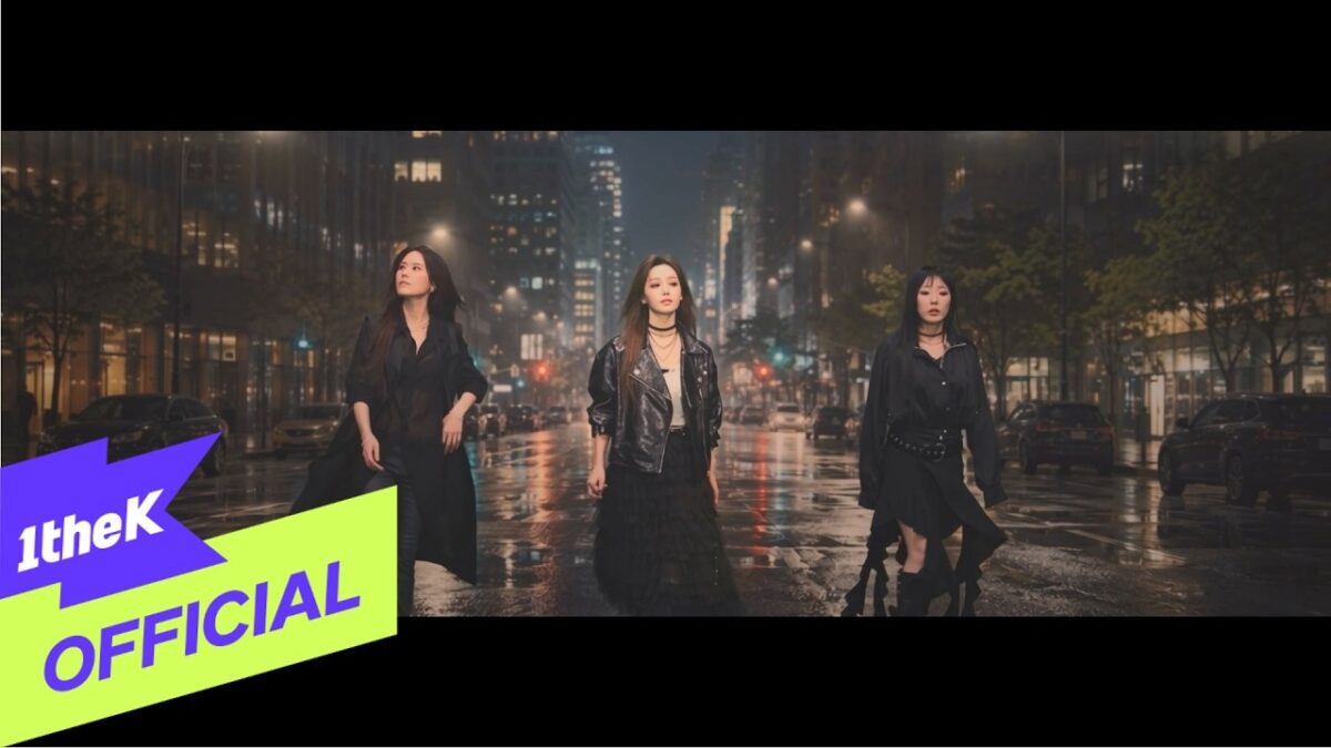 Três mulheres em trajes pretos caminham em uma rua molhada, iluminada por luzes urbanas à noite.