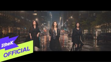 Três mulheres em trajes pretos caminham em uma rua molhada, iluminada por luzes urbanas à noite.