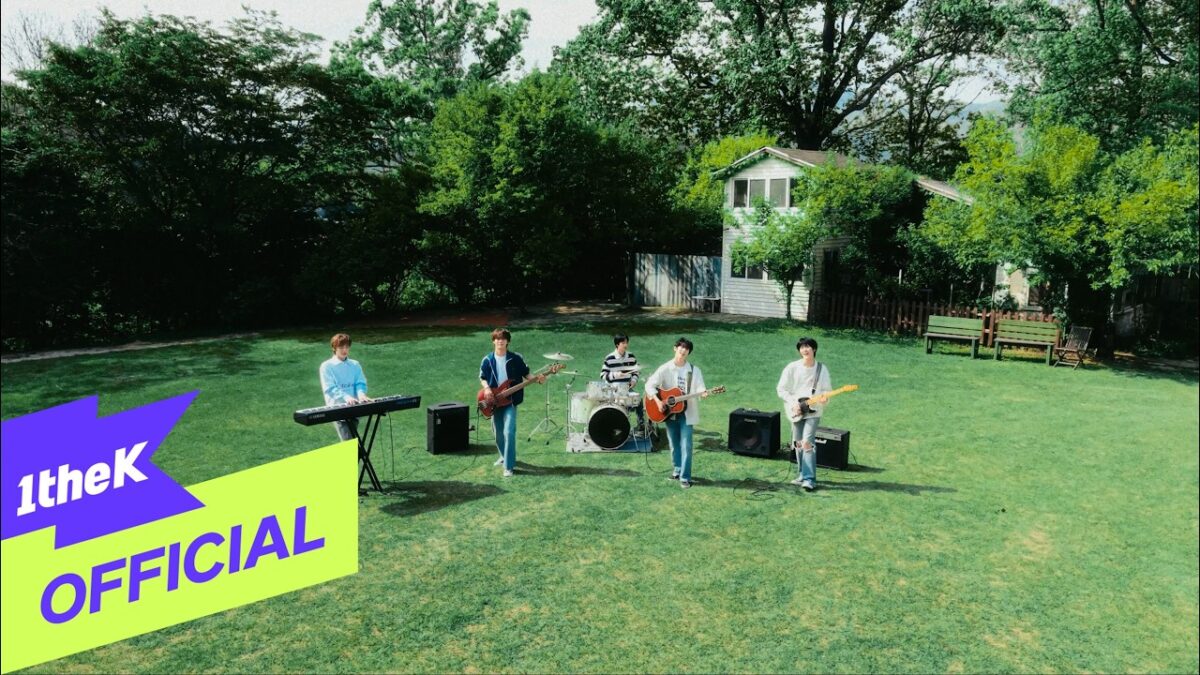 Banda se apresenta ao ar livre, com cinco integrantes tocando instrumentos em um gramado verde e cercados por árvores.