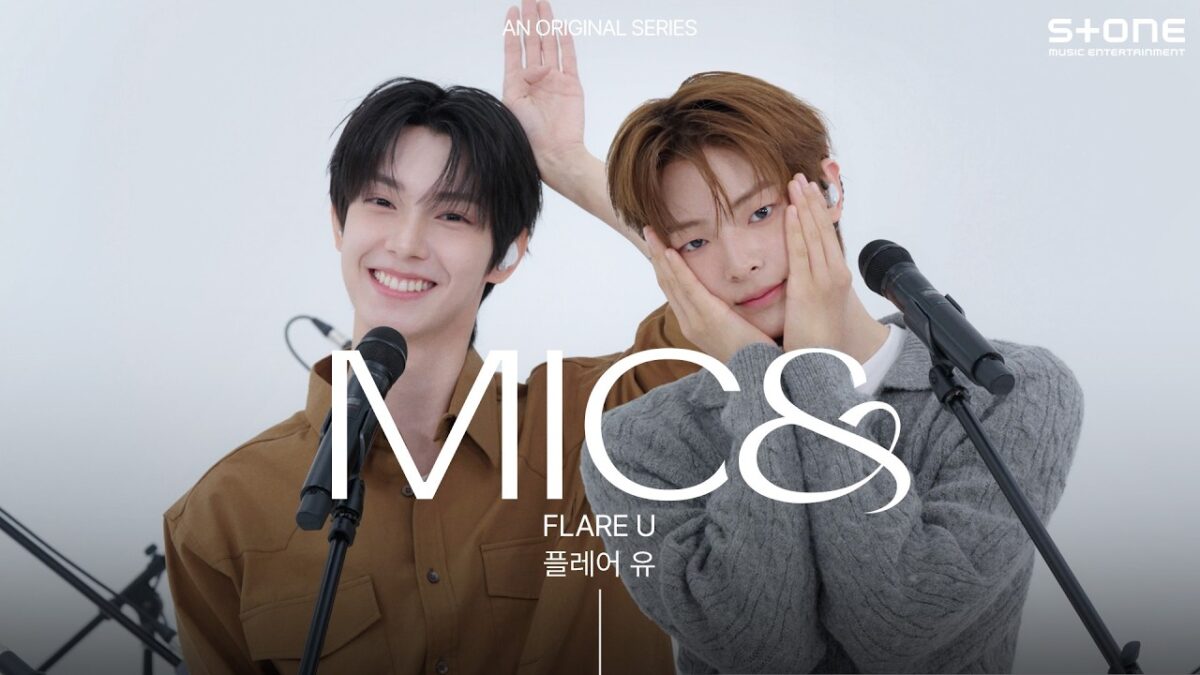 Dois cantores sorrindo em um palco, um deles com as mãos no rosto, prontos para se apresentar ao vivo.
