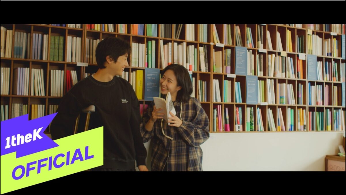 Yun Seong e uma jovem sorridente em uma biblioteca, trocando olhares enquanto seguram um livro juntos.