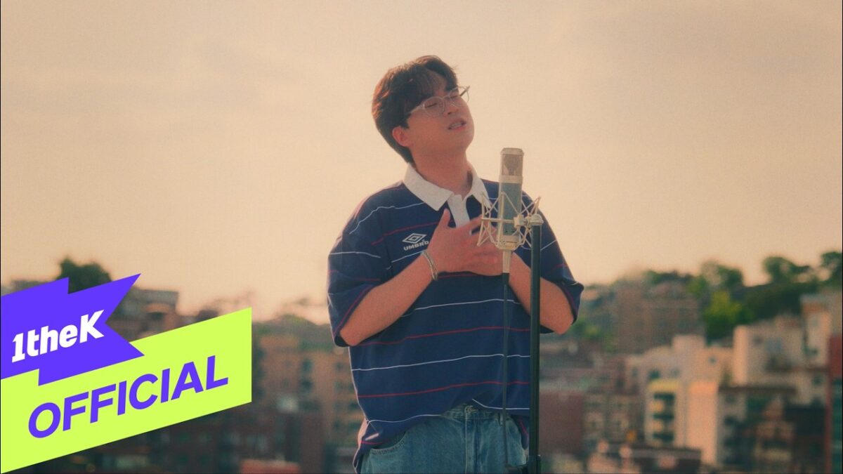Kim Dong Hyun canta emocionado em um telhado, com microfone e vista da cidade ao fundo.