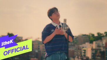 Kim Dong Hyun canta emocionado em um telhado, com microfone e vista da cidade ao fundo.