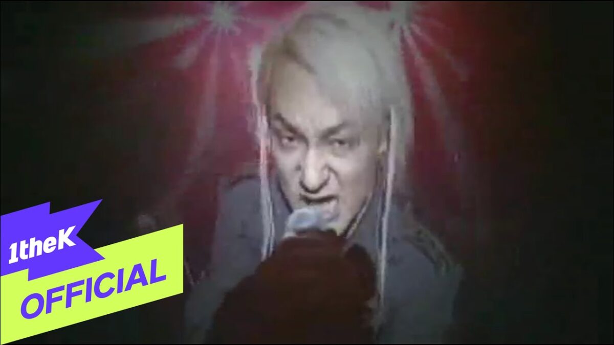Vocalista de cabelo platinado canta intensamente em um videoclipe com luzes vibrantes ao fundo.