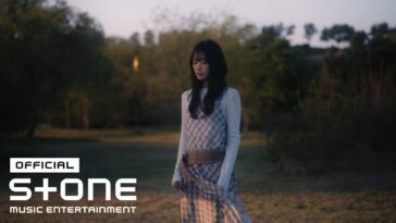 Kim Gyeol canta em um campo ao entardecer, usando um vestido xadrez e segurando um microfone.