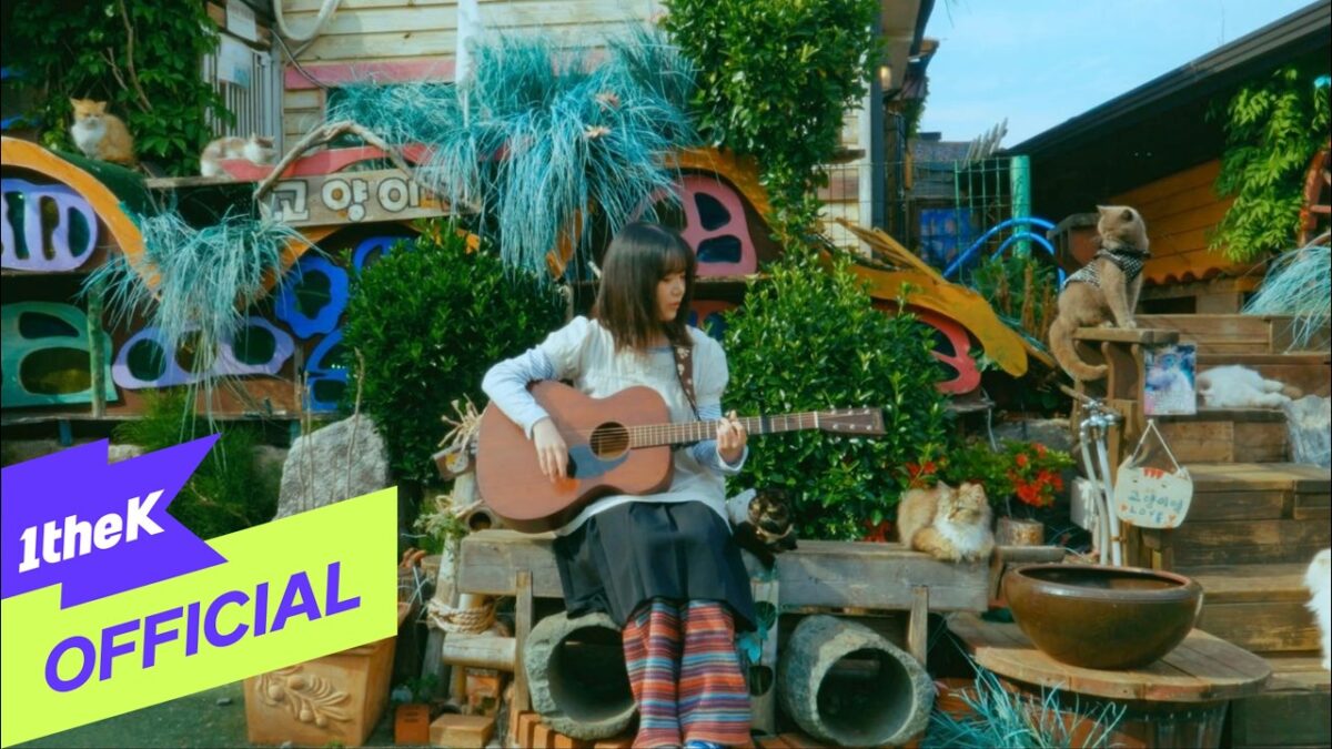 Yeon Seo toca violão em um cenário colorido com gatos e plantas ao fundo.