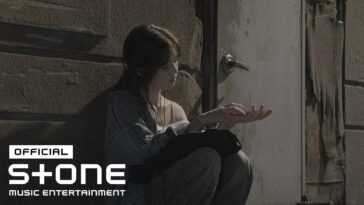Jovem sentada em uma parede, olhando para suas mãos com expressão pensativa, em um ambiente urbano e escuro.