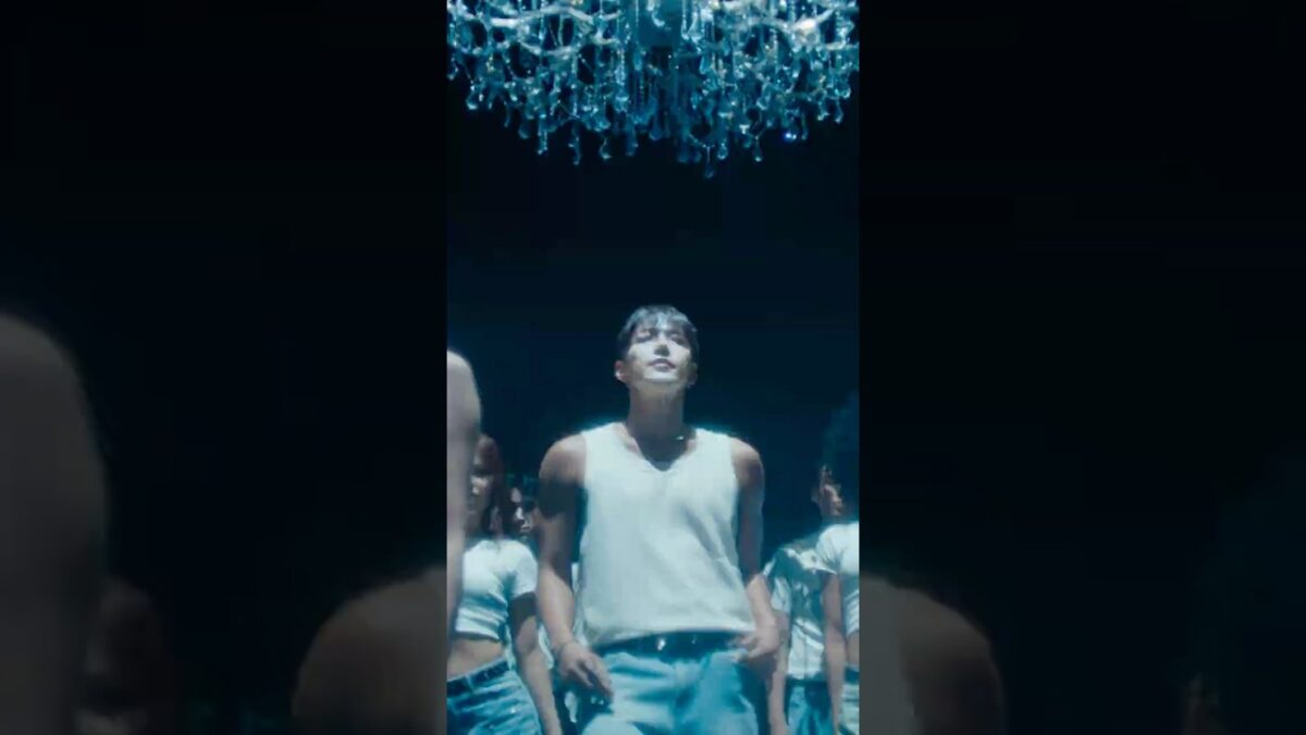 JUN. K em destaque, cercado por dançarinos, sob um lustre brilhante em um cenário escuro.
