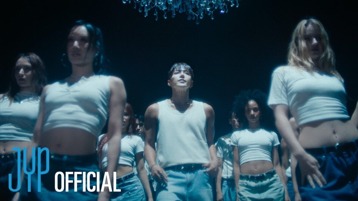 JUN. K e dançarinos em trajes brancos, sob um lustre, criando uma atmosfera intensa e envolvente.