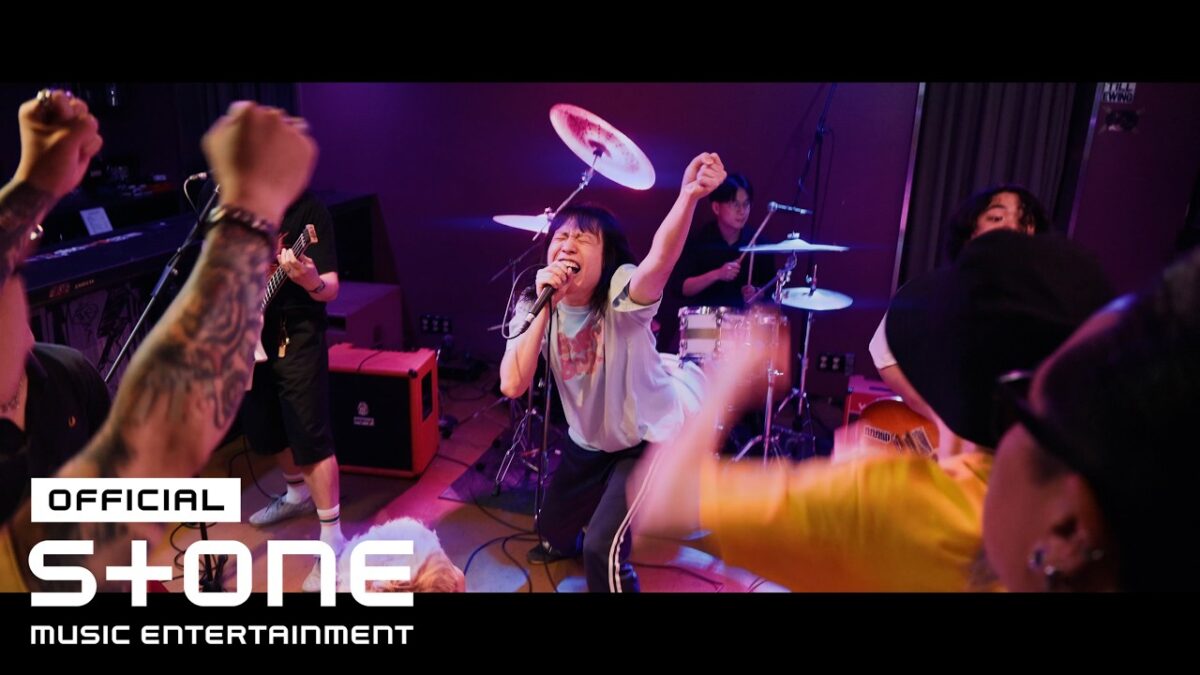 Vocalista performa energicamente no palco, cercado por fãs animados e músicos, durante gravação do clipe "Little Baby".