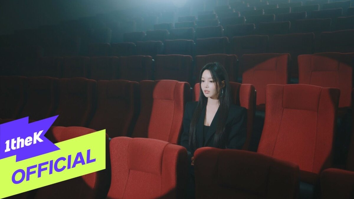 Artista sentada sozinha em uma sala de cinema vazia, iluminada de forma suave.