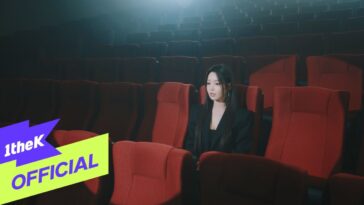 Artista sentada sozinha em uma sala de cinema vazia, iluminada de forma suave.