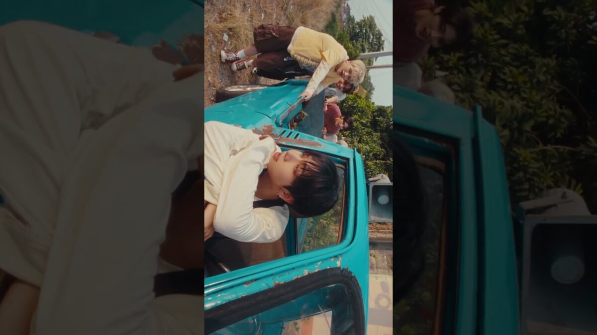 Jovens em um carro azul, um sentado e outros brincando ao fundo, em um cenário natural e descontraído.